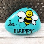 Spread happiness and encouragement! This cute hand-painted rock says Bee Happy and features a yellow and black bumblebee. The background is sky blue. It measures around 3.5 inches in length (size and shape will vary from stone to stone). This stone is painted with acrylics and sealed
