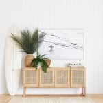 Beautiful beachy vibe with a cane console table in light wood, vertical white shiplap, large wall art, plants, and a surfboard! #entrywaydecor #foyerdecor #modern #coastal