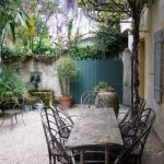 This backyard is adorned with a rustic decor. The dining table is made with a stone design. The chairs look to be made from wood and wicker materials. A green fence is in the background surrounded with an array of plants and trees.